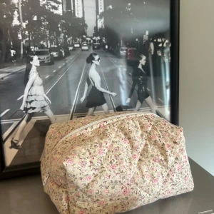 Blommig söt necessär  - Söt quiltad necessär i beige bomullstyg med små blommor i rosa och lila toner. Väskan har en vit dragkedja och ett ljusrosa innerfoder. Perfekt för smink eller småprylar