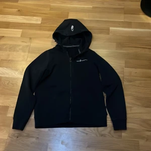 Svart hoodiejacka med dragkedja - Svart hoodiejacka med hel dragkedja och huva. Jackan har en stilren look med diskret logga på bröstet och långa ärmar. Perfekt för dig som gillar en enkel och clean stil. Passar till det mesta och är riktigt skön att ha på sig.
