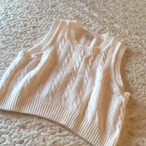 Vit stickad väst från Lindex - Vit kabelstickad väst från Lindex med breda ribbade kanter vid halsringning, ärmhål och nederkant. Klassisk och mjuk modell i stickat material, perfekt att slänga över en skjorta eller t-shirt för en chill look.