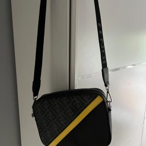 Fendi svart axelväska med gul detalj - Snygg axelväska från Fendi i svart skinn med bred axelrem där Fendi-loggan syns. Framsidan har ett mönstrat parti med Fendi-monogram och en diagonal gul skinnremsa som kontrast. Väskan har dragkedja upptill och stilren, rektangulär form.