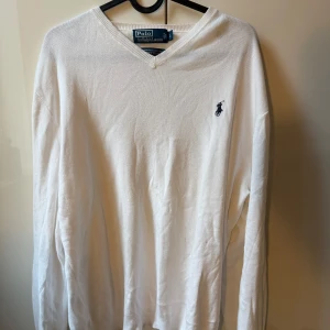 Vit v-ringad tröja Polo Ralph Lauren - Klassisk vit v-ringad tröja från Polo Ralph Lauren med broderad marinblå logga på bröstet. Långärmad och i mjuk bomull, perfekt för en clean och stilren look. Passar dig som gillar tidlös stil och vill ha något som funkar till allt.