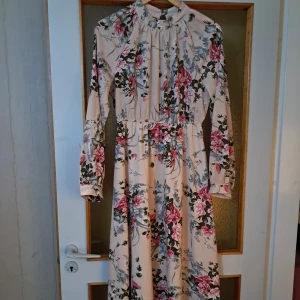 Lång blommig klänning med hög krage - Superfin långklänning i ljusbeige med blommigt mönster i rosa, grönt och blått. Klänningen har hög krage, långa ärmar med manschett och knäppning i nacken. Perfekt för dig som gillar romantisk och feminin stil.