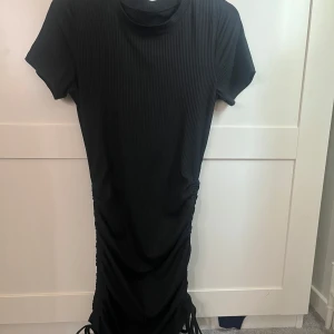 Svart ribbad kortklänning med snörning - Svart ribbad kortklänning med korta ärmar och rund hals. Klänningen har snörningsdetaljer på sidorna nertill som ger en snygg rynkad effekt. Perfekt för dig som gillar en enkel men trendig look.