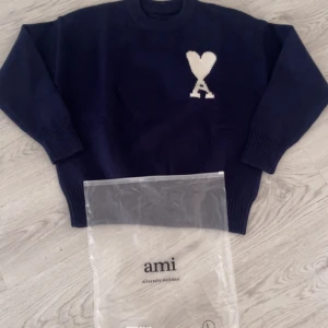 Mörkblå stickad tröja från Ami - Snygg mörkblå stickad tröja från Ami med vit hjärtan-A logga på bröstet. Tröjan har rund hals, ribbade muddar och är tillverkad i 100% ull. Perfekt för dig som gillar stilrena detaljer och vill ha något som sticker ut lite extra. Hör av er vid funderingar eller intresse❤️💞‼️✅🚌🥺