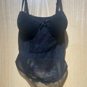 Svart spetskorsett med rosett - Snygg svart korsett med spetsdetaljer och meshpaneler. Kuporna har ett coolt bikakemönster och framtill sitter en liten söt rosett. Justerbara axelband och hakknäppning bak. Perfekt för att lyfta din outfit med en edgy vibe. Aldrig använd! 