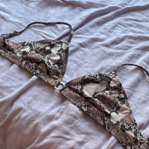 Bikinitopp med ormmönster och gulddetalj - Säljer en trekantsformad bikinitopp med trendigt ormmönster i beige, brunt och svart. Toppen har smala axelband och en snygg guldfärgad spänndetalj bak. Perfekt för dig som vill sticka ut på stranden i sommar!