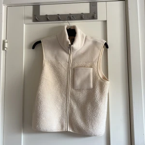 Beige fleeceväst från Pieces - Mysig beige fleeceväst från Pieces med hög krage och dragkedja framtill. Västen har en bröstficka och är tillverkad i mjukt teddyfleece-material. Perfekt att slänga över en hoodie eller långärmad tröja för extra värme.