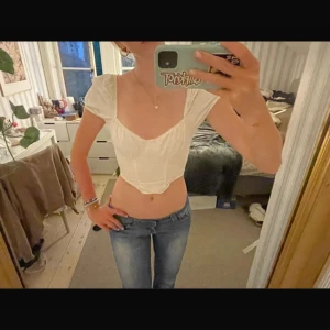 Vit croppad korsett-topp - Supersnygg vit korsett-topp med korta puffärmar och markerad byst. Toppen är croppad med spetsig nederkant och har en romantisk vibe. Perfekt att styla med jeans eller kjol för en trendig look.