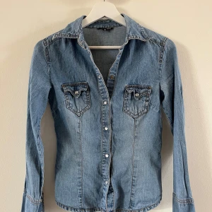 Klassisk jeansskjorta med knappar - Snygg ljusblå jeansskjorta med långa ärmar och två bröstfickor med knappdetaljer. Skjortan har en figurnära passform och klassisk krage. Perfekt att styla till både vardagliga och trendiga outfits.