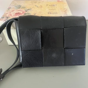 Svart axelväska i flätad skinn - Säljer en svart axelväska i skinn med flätad design. Väskan har en stilren och modern look med bred axelrem och fyrkantiga paneler. Perfekt för dig som vill ha en snygg accessoar till vardags.