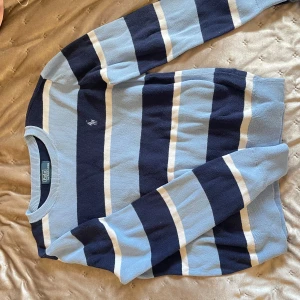 Randig tröja från Polo Ralph Lauren - Säljer en snygg långärmad stickad tröja från Polo Ralph Lauren i blått, mörkblått och vitt. Tröjan har breda ränder och en broderad logga på bröstet. Den är i storlek 10-12 år från Kidsbrandstore