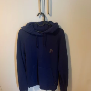 Mörkblå hoodie med broderad detalj - Säljer en mörkblå hoodie med huva. Tröjan har en liten broderad detalj på bröstet och en stor ficka framtill. Perfekt för dig som gillar en avslappnad stil. 