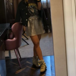 Silverfärgad kortkjol med resårmidja - Säljer en glansig silverfärgad kortkjol med bred resår i midjan. Kjolen har en lätt A-linjeform och är tillverkad i ett syntetmaterial som ger en skimrande effekt. Perfekt för dig som vill sticka ut lite extra eller till tema fest space eller liknande!