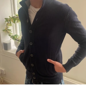 Svart stickad cardigan med knappar - Mysig mörkblå stickad cardigan med grov ribbad krage och knäppning framtill. Jackan har långa ärmar och breda muddar vid ärmslut och nederkant. Perfekt för lager-på-lager och en avslappnad stil.