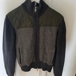 Grå stickad jacka med gröna detaljer från Moncler - Snygg grå stickad jacka från Moncler med quiltade gröna axelpartier och armbågslappar. Jackan har dragkedja framtill, höga muddar och två fickor med dragkedja. Perfekt för kyliga dagar och har en modern, sportig look.