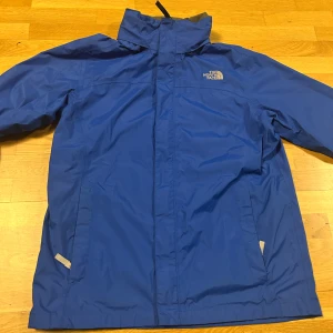 Blå vindjacka från The North Face - Säljer en blå vindjacka från The North Face med hög krage och dragkedja framtill. Jackan har två sidofickor och meshfoder på insidan för extra komfort. Perfekt för blåsiga dagar och enkel att matcha med olika outfits. Står XL men passar S/Xs