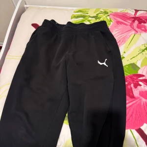 Svarta mjukisbyxor från Puma - Svarta mjukisbyxor från Puma med resår i midjan och vit Puma-logga på ena benet. Perfekta för en avslappnad stil eller träning. Byxorna har en klassisk passform och är gjorda i mjukt material.