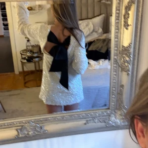 Vit paljettklänning med öppen rygg och rosett - Säljer en superfin vit paljettklänning med lång ärm och stor svart rosett i ryggen. Klänningen har öppen rygg och är täckt av glittrande paljetter, perfekt för att sticka ut. Kort modell med en elegant och festlig känsla. Hm populär