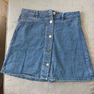 Jeanskjol med knappar från Noisy May - Säljer en klassisk blå jeanskjol från Noisy May i medium. Kjolen har knappar framtill, fickor både fram och bak men bara dem där bak funkar att öppna. A-linjeformad och i mjukt denimtyg, 
