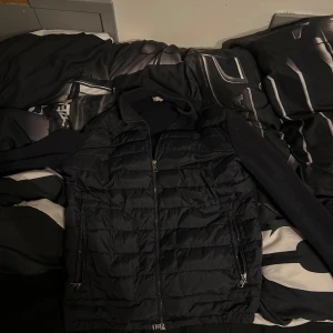 Svart dunväst med ärmar och dragkedja - Snygg svart väst med vadderad framsida och ärmar i mjukt material. Västen har dragkedja framtill och två fickor med dragkedja. Perfekt för lager-på-lager-stil.