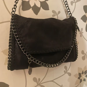Svart axelväska med kedjedetaljer - Snygg svart axelväska i mocka-look med grova kedjor längs kanterna och som axelrem. Väskan har lockstängning och insida med leopardmönstrat foder samt innerfack med dragkedja. Perfekt för dig som vill ha en inte för stor och inter för liten handväska!