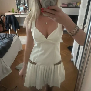 Vit plisserad kortklänning från Zara - Säljer en vit kortklänning från Zara med djup v-ringning och plisserad kjol. Klänningen har smala axelband och dekorativa bältesdetaljer i midjan. Perfekt för sommarens alla tillfällen!