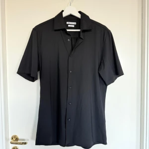 Svart kortärmad skjorta från Zara - Säljer en svart kortärmad skjorta från Zara i slim fit-modell. Skjortan har klassisk krage och knäppning framtill. Perfekt för dig som gillar en stilren och enkel look. Materialet känns mjukt och lätt. Perfekt till sommaren.