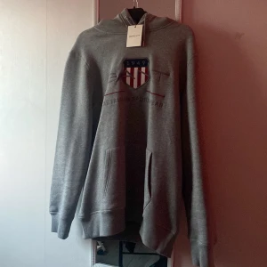 Grå hoodie från GANT med broderad logga - Säljer en grå hoodie från GANT med broderad logga och sköld på bröstet. Tröjan har känguruficka och huva med dragsko. Perfekt för en avslappnad och sportig stil.