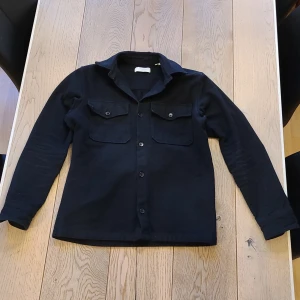 Svart overshirt från Jack & Jones - Stilren svart overshirt från Jack & Jones med klassisk krage och två bröstfickor med knappar. Skjortan har långa ärmar och stängs med knappar framtill. Perfekt att bära som lager över en t-shirt.