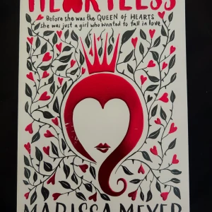 Heartless - En fantasifull roman av Marissa Meyer som berättar historien om Hjärter Dam innan hon blev drottning. Omslaget är vitt med svarta och röda detaljer, hjärtan och en krona. Perfekt för dig som gillar sagoinspirerade berättelser och romantik.