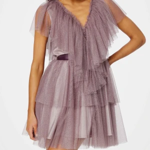 Ida Sjöstedt Lila glittrig klänning - Superfin lila klänning med glittrigt tylltyg. Klänningen har v-ringning framtill, djupare v-ringning i ryggen, korta volangärmar och ett matchande satinband i midjan. Perfekt för speciella tillfällen! Nypris 4 999kr.