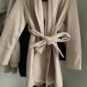 Beige kappa med bälte - Säljer en stilren beige kappa med bälte i midjan och långa ärmar. Jackan har en klassisk krage och är enkel att matcha till olika outfits. Perfekt för dig som gillar en tidlös look.