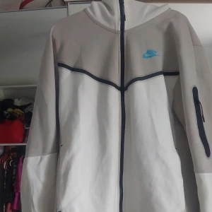 Vit och grå Nike tech med huva - !PRIS KAN DISKUTERAS! Säljer en stilren vit och ljusgrå Nike tech med svart dragkedja och blå Nike-logga på bröstet. Jackan har huva, lång ärm med ficka på ena ärmen och sportig design. Perfekt för dig som gillar streetwear och sportig stil. Storlek s och använd 3-2 gånger
