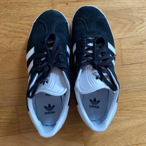 Adidas Gazelle svartvita sneakers - Säljer ett par klassiska Adidas Gazelle sneakers i svart mocka med vita detaljer och de ikoniska tre ränderna på sidan. Skorna har snörning och vit sula samt Adidas-logga på häl och plös. Perfekta för en stilren vardagslook. Använda 2 gånger.