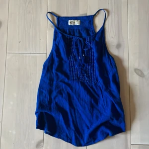 Kornblå linnetopp från Hollister - Säljer en kornblå linnetopp från Hollister med smala axelband och knytning framtill. Toppen har dekorativa sömmar 🪩🪽säljer pågrund av för stort 