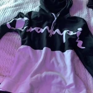 Svart och vit hoodie från Champion - Säljer en snygg hoodie från Champion i svart och vitt med stor logga över bröstet. Tröjan har huva med dragsko och är tillverkad i mjukt bomullsmaterial. Perfekt för en avslappnad och sportig stil. funkar för alla! båda snörena finns kvar så man kan välja själv Lite fläckar på magen fram men syns inte jättemycket och lätt att få bort