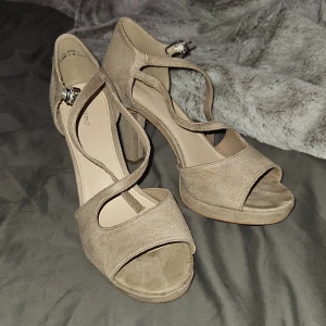 Beige sandaler med klack och öppen tå - Säljer ett par stilrena beige sandaler med klack och öppen tå. Skorna har en snygg korsad rem över foten och justerbart spänne vid ankeln. Perfekta för dig som vill ha en elegant men enkel look.