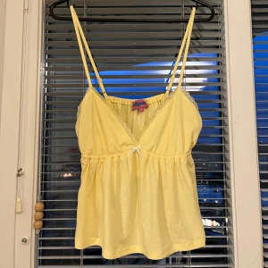 Gul linnetopp med spetsdetaljer - Söt gul linnetopp med tunna axelband och spetsdetaljer längs urringningen. Toppen har en liten rosett framtill och är lätt rynkad under bysten för en feminin siluett. Perfekt för varma dagar.