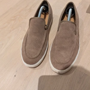 Bruna loafers i stl 42 - Säljer ett par bruna loafers sneakers i mocka med perforerat mönster och vit platt sula. Skorna har rund tå och är perfekta för en avslappnad stil. Enkla att ta på och av tack vare slip-on designen.