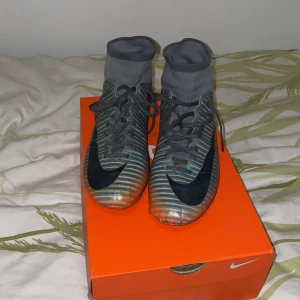 Nike Mercurial CR7 fotbollsskor - Säljer ett par Nike Mercurial CR7 fotbollsskor i grått med svarta detaljer och orange sula. Skorna har hög strumpkrage för extra stöd och är designade för spel på gräs. Perfekta för dig som vill ha stil och prestanda på planen.( one of a kind) Kommer inte med boxen som är på bilden!!!