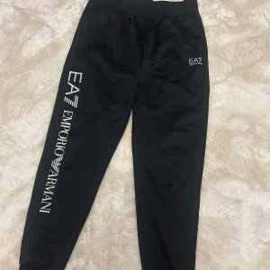 Svarta mjukisbyxor från Emporio Armani EA7 - Svarta mjukisbyxor från Emporio Armani EA7 med stort vitt tryck längs ena benet och mindre logga vid fickan. Byxorna har resår i midjan och är perfekta för en avslappnad stil.