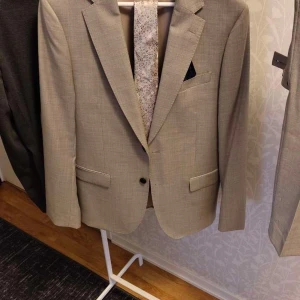 Beige kostym från Dressmann i ull - Säljer en stilren beige kostym från Dressmann i 100% ull. Kavajen har två knappar, klassiska slag och bröstficka med näsduk. Byxorna har bälteshällor och rak passform. Perfekt för dig som vill ha en tidlös look.Pris går att diskutera❤️