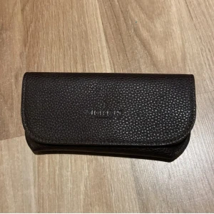 Svart glasögonfodral från Morris - Snyggt svart glasögonfodral från Morris i präglat läder. Fodralet har en stilren design med diskret logotyp på framsidan och insidan. Perfekt för att skydda dina glasögon med lite extra stil.