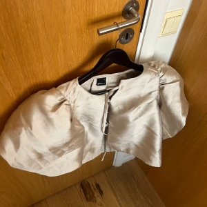 Guldig topp med puffärmar  - Säljer en beige topp med puffärm från gina med stora puffärmar och öppen front med knytning. . Perfekt för att lyfta en outfit med något unikt och trendigt.