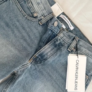 Blå raka jeans från Calvin Klein Jeans - Snygga blå jeans från Calvin Klein Jeans med raka ben och slitna detaljer på båda benen. Klassisk femficksmodell med normal passform och hög midja. Perfekta för en avslappnad och trendig look.