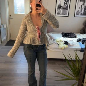 Gråvit stickad kofta med rosett från Nelly - Superfin gråvit stickad kofta från Nelly med mönster och en rosa rosett framtill. Koftan har lång ärm, peplumdetalj nedtill och ett klassiskt nordiskt mönster. Perfekt för lager på lager och en mysig look.