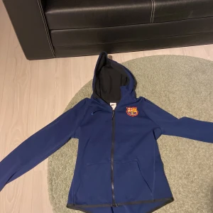 Blå Nike FC Barcelona hoodie med dragkedja - Säljer en blå hoodie från Nike med FC Barcelona-logga på bröstet. Jackan har huva, dragkedja framtill och två fickor. Perfekt för dig som älskar fotboll och vill representera ditt lag.