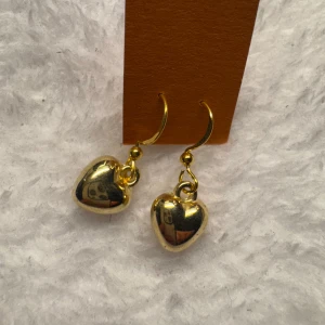 Guldiga hjärtformade örhängen - Säljer ett par glansiga örhängen i guld med hängande hjärtan. Perfekta för att ge en söt och trendig touch till din stil. Enkla att matcha med olika outfits och passar dig som gillar detaljer. Krokarna är i sterling silver