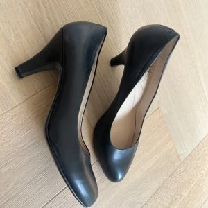 Svarta pumps med klack Vaga Bond - Klassiska svarta pumps med rund tå och medelhög klack. Skorna har en stilren design och är tillverkade i ett slätt material som ger en elegant look. Perfekta för dig som vill ha ett par tidlösa och mångsidiga klackar.