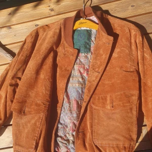 Brun mockajacka med mönstrat innerfoder  - Brun mocka/suede jacka med resårband i midjan, kragen och vid handlederna. Jackan har två fickor fram och en vacker innerfoder. Har aldrig använts av mig och är i väldigt bra skick.